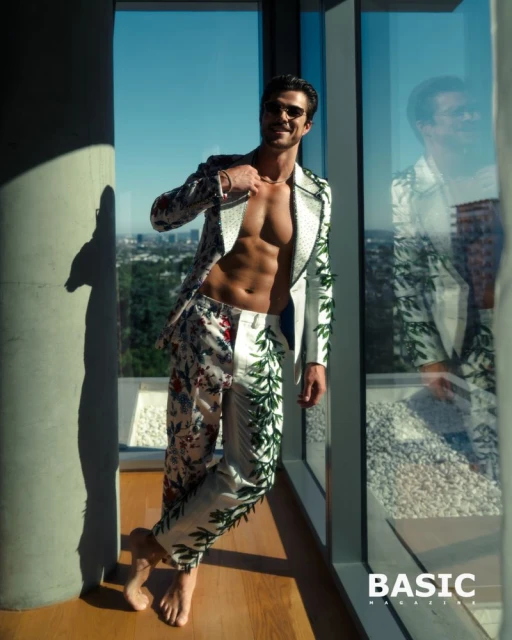 Athletic male model with open floral suit poses by window in a fashion shot for Basic Magazine, showcasing abs, chest and fashionable suit.