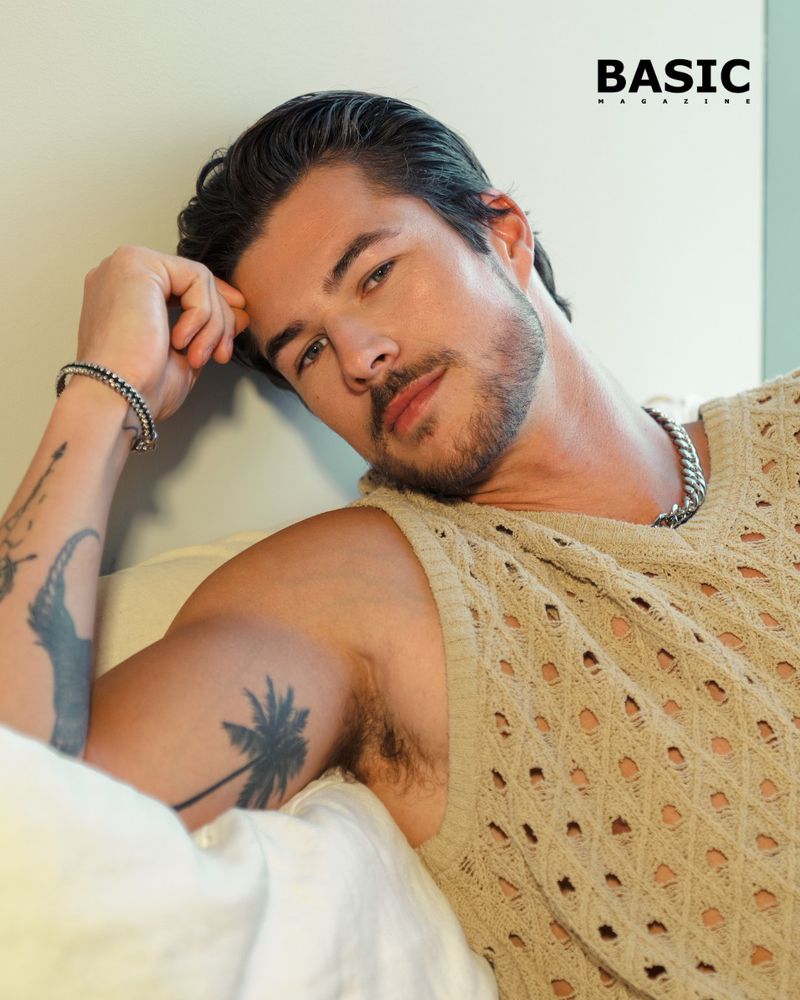 Chase Mattson wearing an open-knit beige sleeveless top and silver chain necklace, leaning against white pillows.