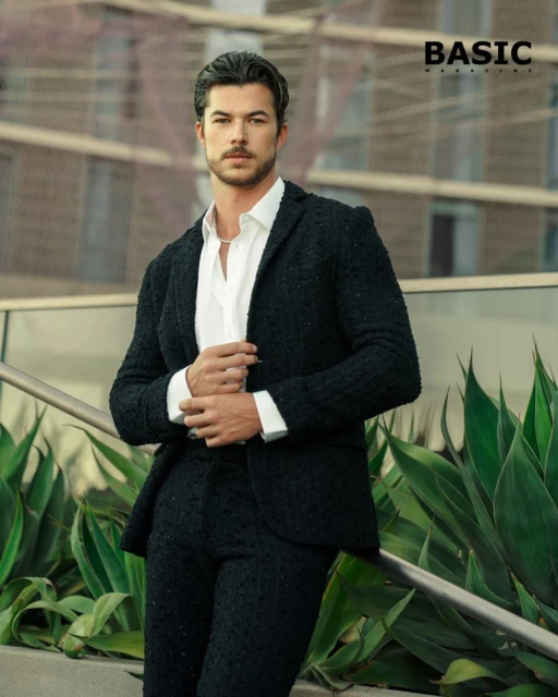 Man in a black tweed suit stands outdoors next to a staircase and succulent plants, wearing a white shirt in front of a building.