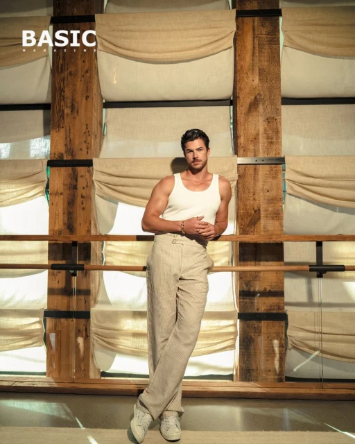 Portrait of a male model in casual wear leaning against a wooden structure with neutral-toned drapery in the background, fashion editorial.