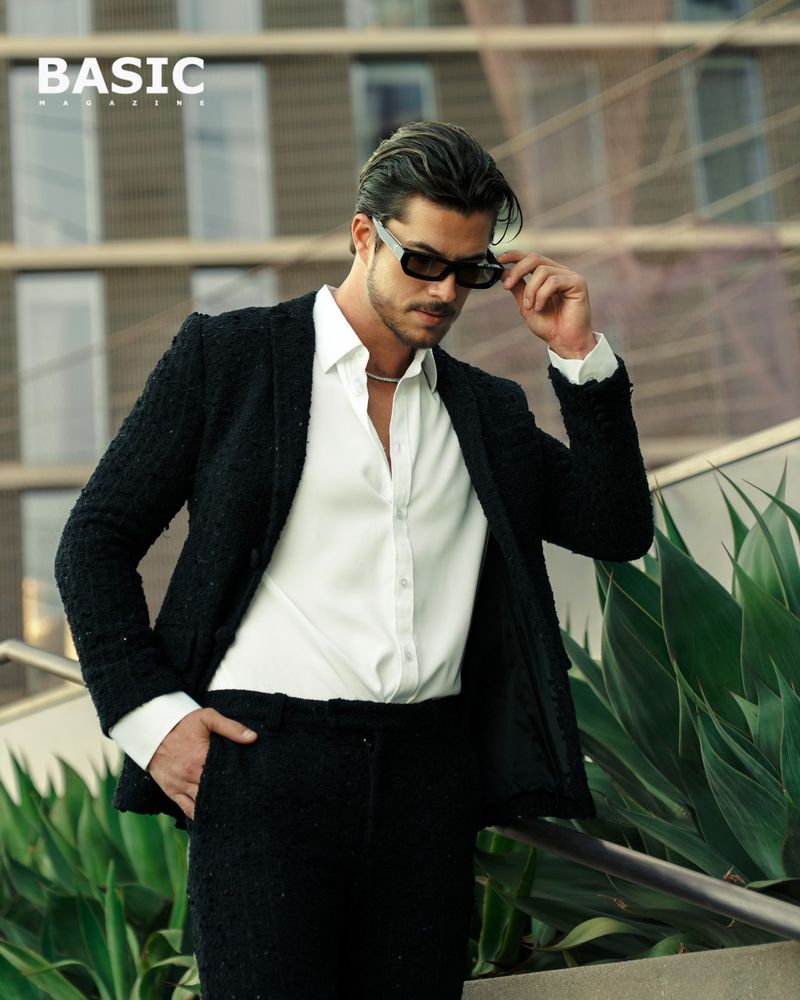 Man in black suit and sunglasses stands outdoors with modern architecture and green plants visible; fashion editorial style.