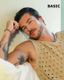 Chase Mattson wearing an open-knit beige sleeveless top and silver chain necklace, leaning against white pillows.
