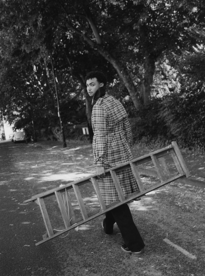 Monochrome portrait of person in plaid coat carrying a wooden ladder on a paved road, surrounded by trees and greenery.