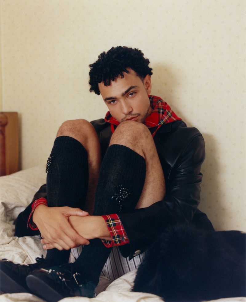 Portrait of a young adult male sitting on a bed with legs crossed, wearing a black jacket, tartan accents, black socks, and black shoes.