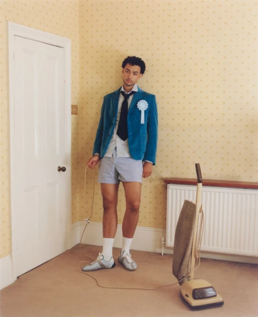 Man in blazer and shorts stands indoors with vintage vacuum. He wears a rosette. The room is carpeted with patterned wallpaper.