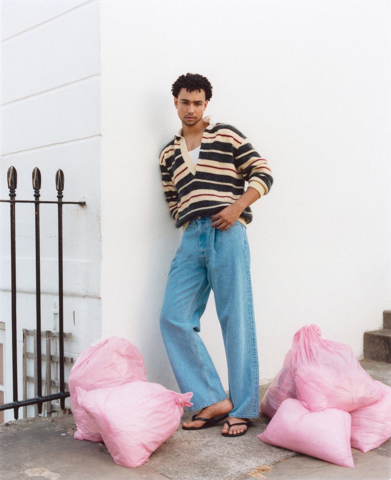 Man poses in jeans and striped sweater, leaning against white wall. Pink bags on ground add a striking contrast to the minimalist setting.