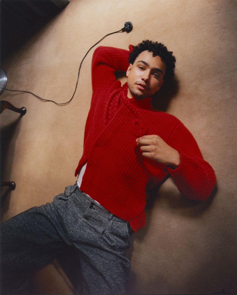 Young adult male fashion portrait: Man lying on carpet in red sweater and grey pants, looking at camera with neutral expression.