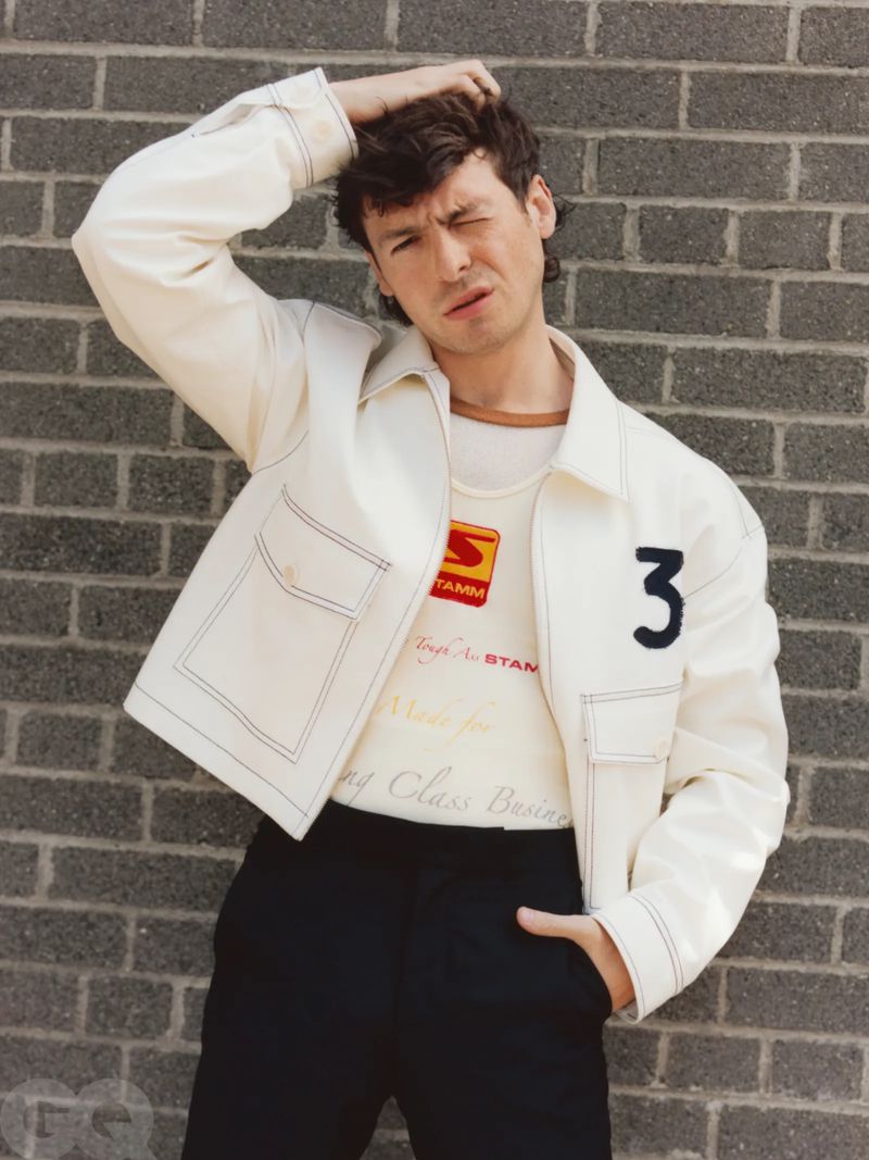 Young adult male poses in front of a brick wall, wearing a white jacket and black pants in a medium shot with eye-level angle.