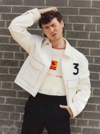 Young adult male poses in front of a brick wall, wearing a white jacket and black pants in a medium shot with eye-level angle.