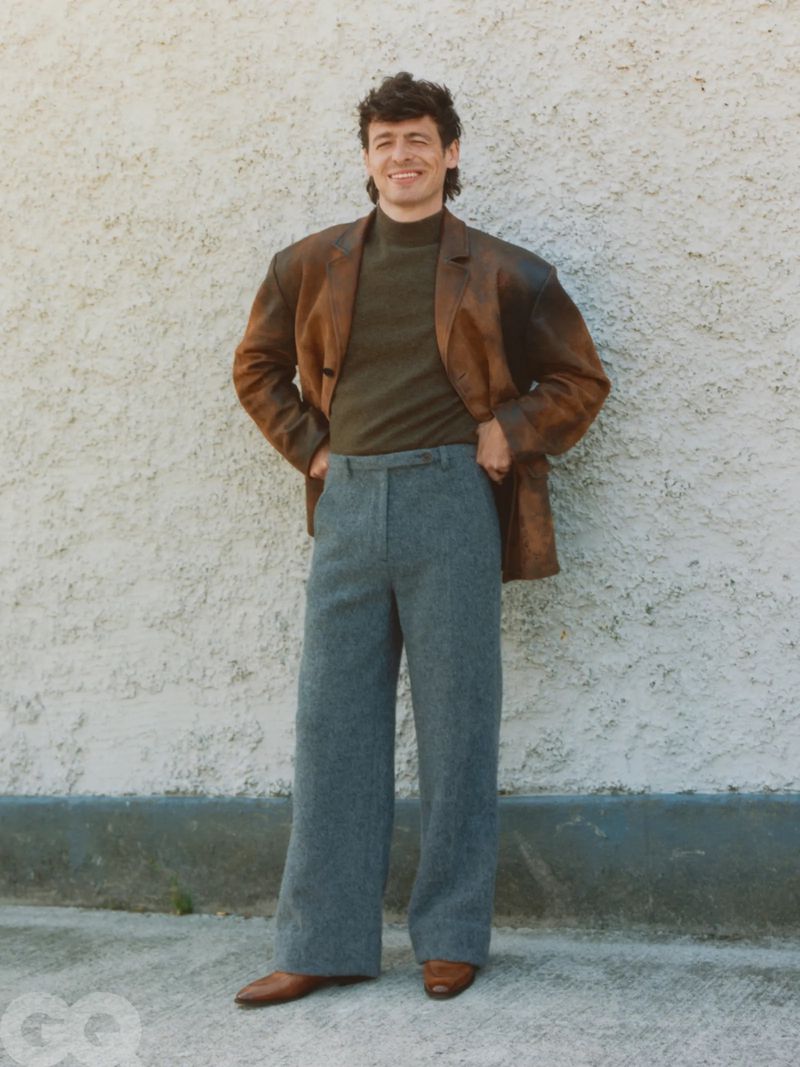 An adult male stands smiling outdoors wearing a brown jacket, olive green turtleneck, and grey trousers, with a textured wall background.