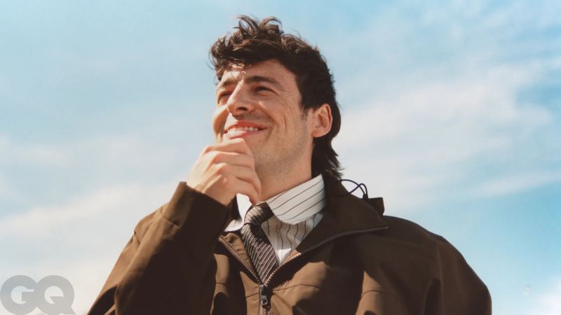 Anthony Boyle smiling outdoors against a clear blue sky wearing a brown jacket, striped shirt, and tie for British GQ.