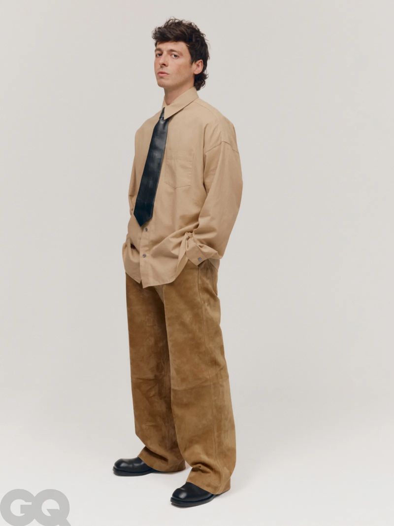 Studio shot of a young adult male posing in tan oversized shirt and wide-legged brown pants, with a black tie and black shoes.