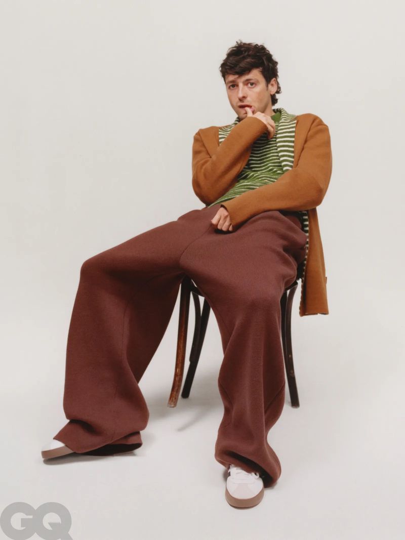 Adult male in oversized brown trousers and striped top posing in a photo studio while sitting on a chair.