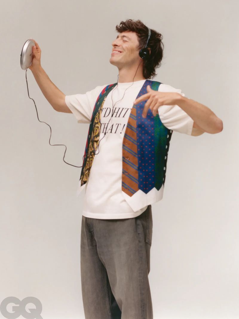A man wearing a vest and headphones dances while holding a CD player in a studio with a light gray background, casual but fashionable.