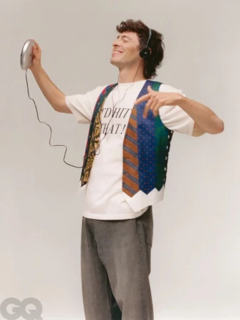 A man wearing a vest and headphones dances while holding a CD player in a studio with a light gray background, casual but fashionable.