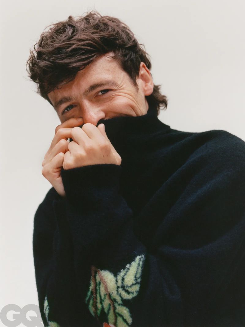 Close-up studio portrait of an adult male with curly dark hair, wearing a floral-patterned turtleneck sweater against a neutral backdrop.
