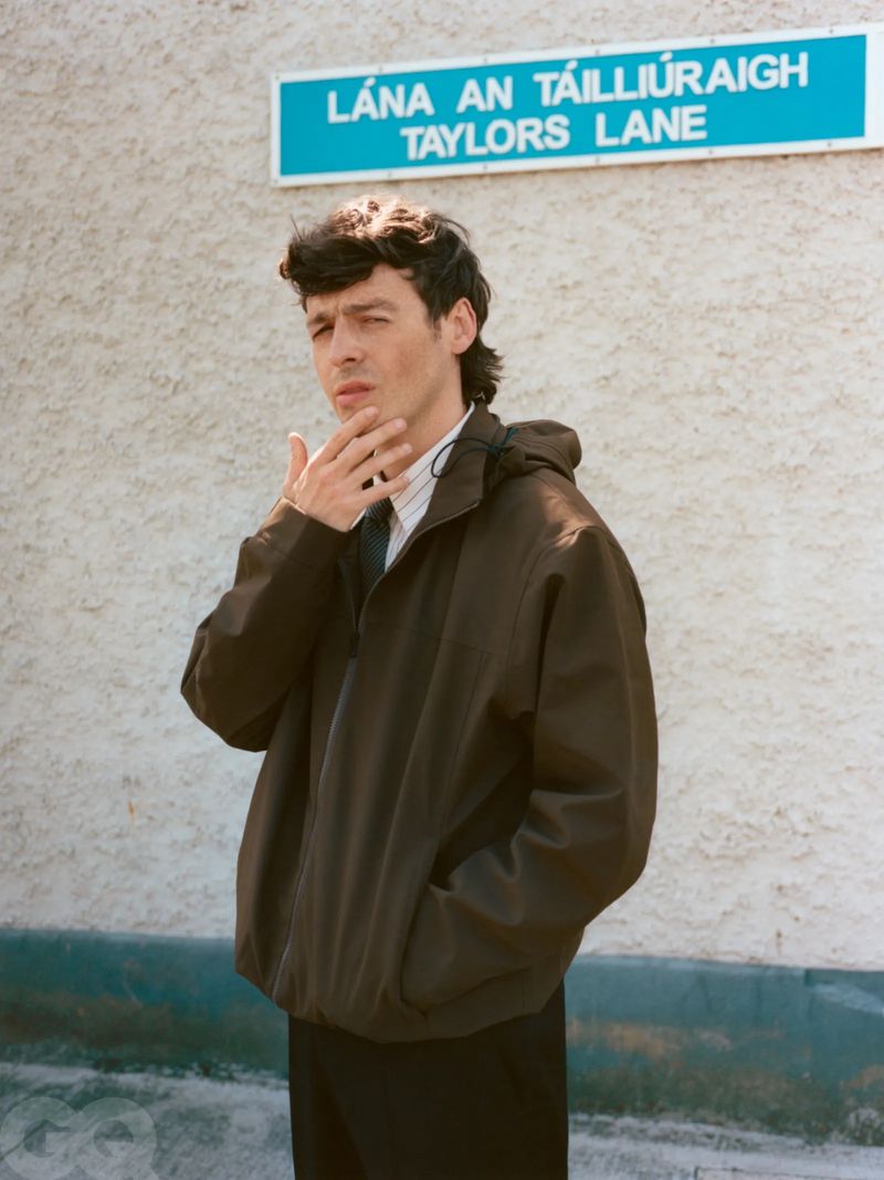Adult male in a brown jacket, standing near a blue street sign on a textured grey wall, holds a hand to his face in an outdoor setting.