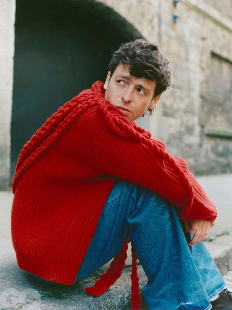 Portrait of a young adult male with dark hair wearing a red knit sweater and jeans, seated against a stone wall. GQ logo in corner.