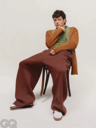 Anthony Boyle seated on a dark wooden chair wearing a brown cardigan, green striped sweater, and wide brown trousers against a white background