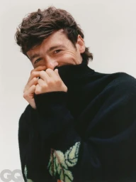 Actor Anthony Boyle wearing a black turtleneck sweater, smiling with hands covering part of his face against a plain light background