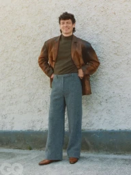 Anthony Boyle wearing a brown leather jacket and grey trousers standing against a textured wall for British GQ