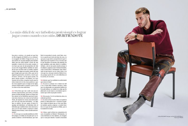A man with fair skin sits on a chair wearing a dark red sweater, patchwork leather pants, and grey boots against a white background.