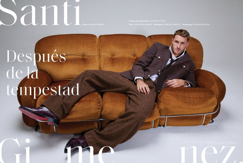 A blond man in a brown suit reclines on a rust-colored velvet sofa, wearing dress shoes, with magazine text visible on a grey background.