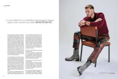 A man in a burgundy sweater and multi-colored leather trousers sitting on an orange chair against a white studio background.