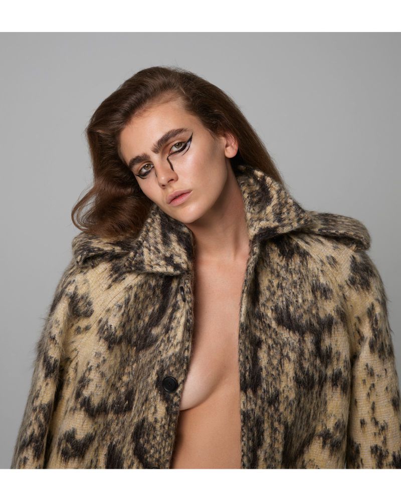A woman with graphic eyeliner posing in a studio, wearing a patterned coat. The image features a neutral background and soft studio lighting.
