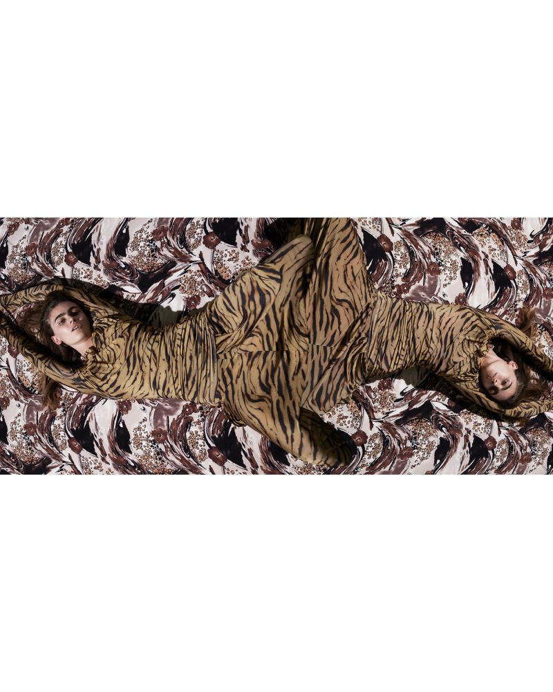 Two women lie side-by-side in matching tan tiger-striped bodysuits on a patterned fabric backdrop in a studio fashion editorial shot.