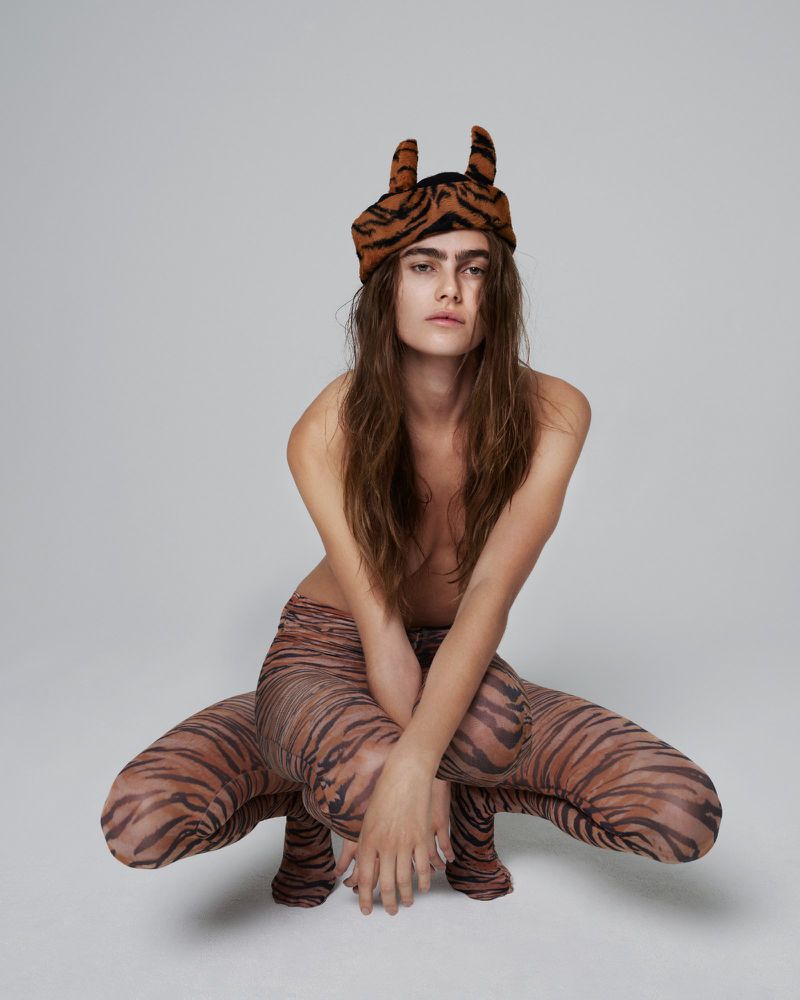 Model poses in studio wearing a tiger-print hat and tights. The individual is crouched with their arms crossed and looking at the camera.