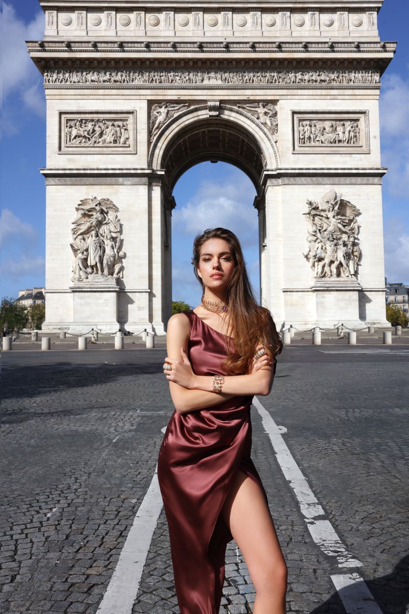 Woman in a burgundy dress posing in front of the Arc de Triomphe in Paris, France, with cobblestone streets and clear sky visible.