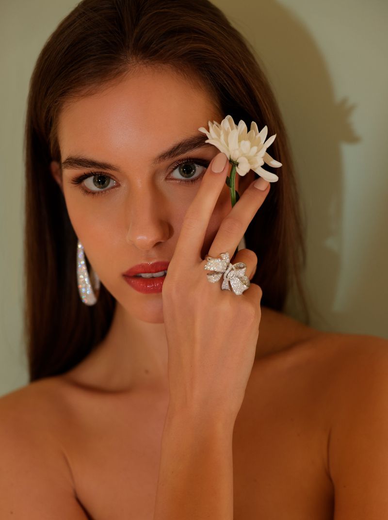 Portrait of a young woman with dark hair holding a white flower, wearing large sparkly hoop earrings and a bow ring on her hand.