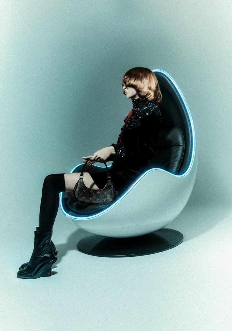 A woman with auburn hair sits in an egg chair with neon trim, dressed in black with a patterned bag and boots against a pale blue background.