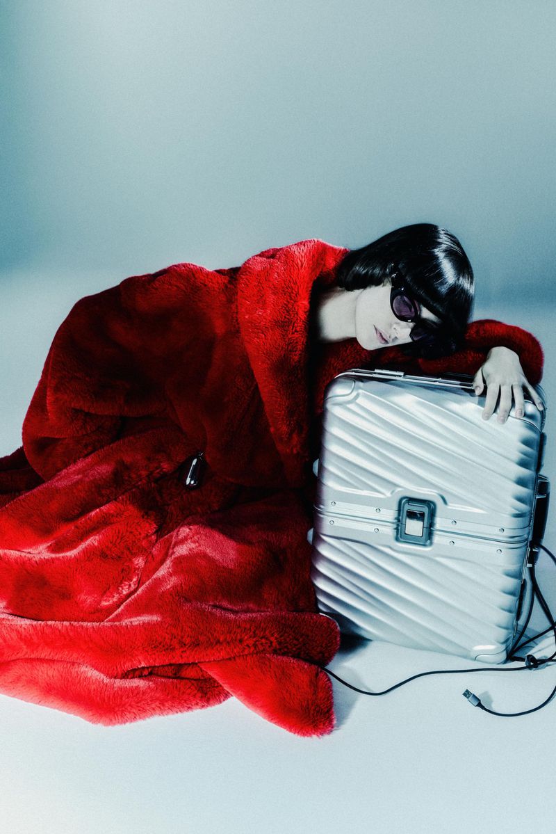 Adult female poses in a red faux fur coat next to a silver suitcase on a light-blue background in a fashion-style product studio shot.