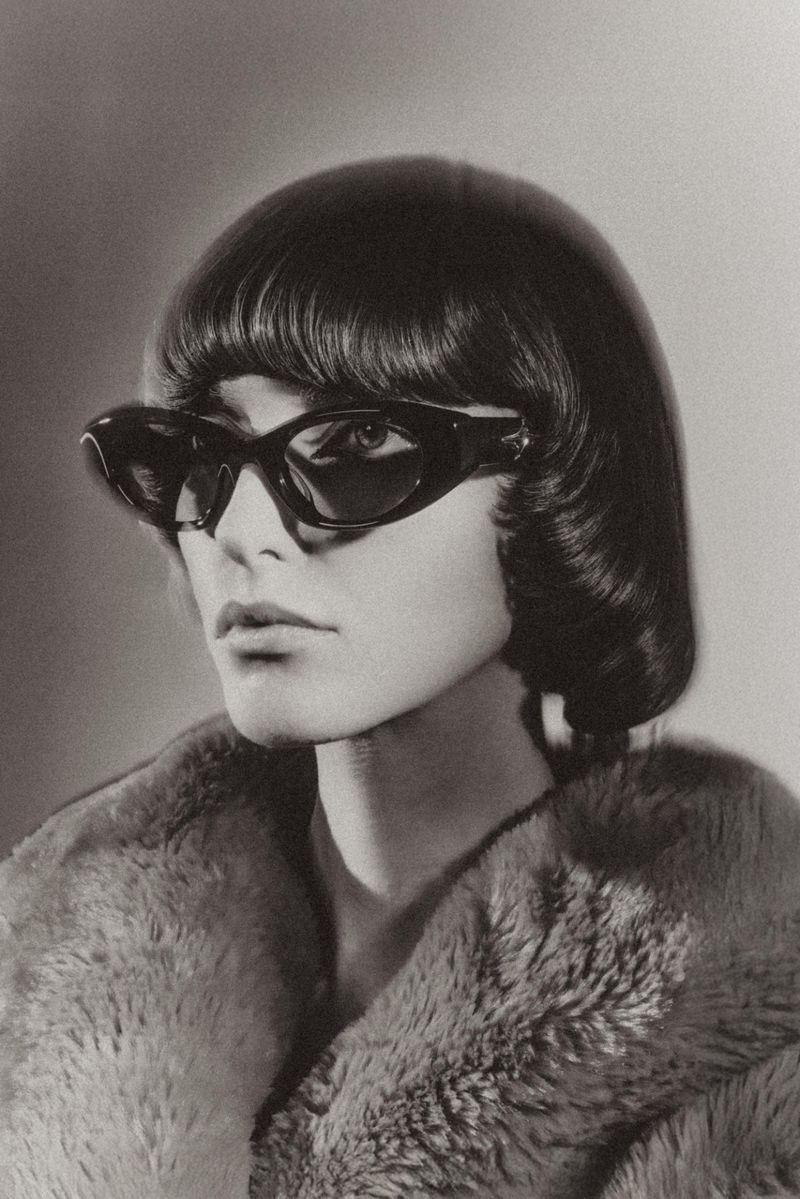 Model wearing dark sunglasses and a textured fur coat with a short bob haircut against a gradient background.