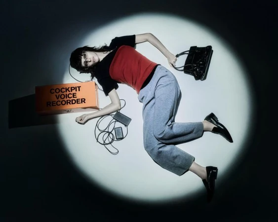 Fashion shot of a woman in grey pants, black top and black loafers, posed in a spotlight with orange box and black purse.