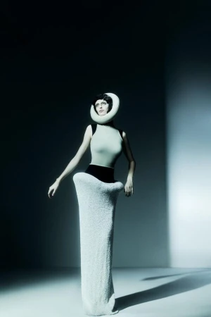 Woman in a light green bodysuit and white avant-garde dress with a circular neckpiece posing in a dimly lit, neutral studio setting.