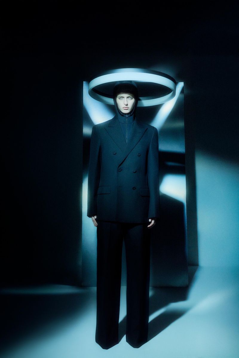 Fashion model in a black suit stands in a dark studio with a ring light overhead, creating a modern and stylish atmosphere.
