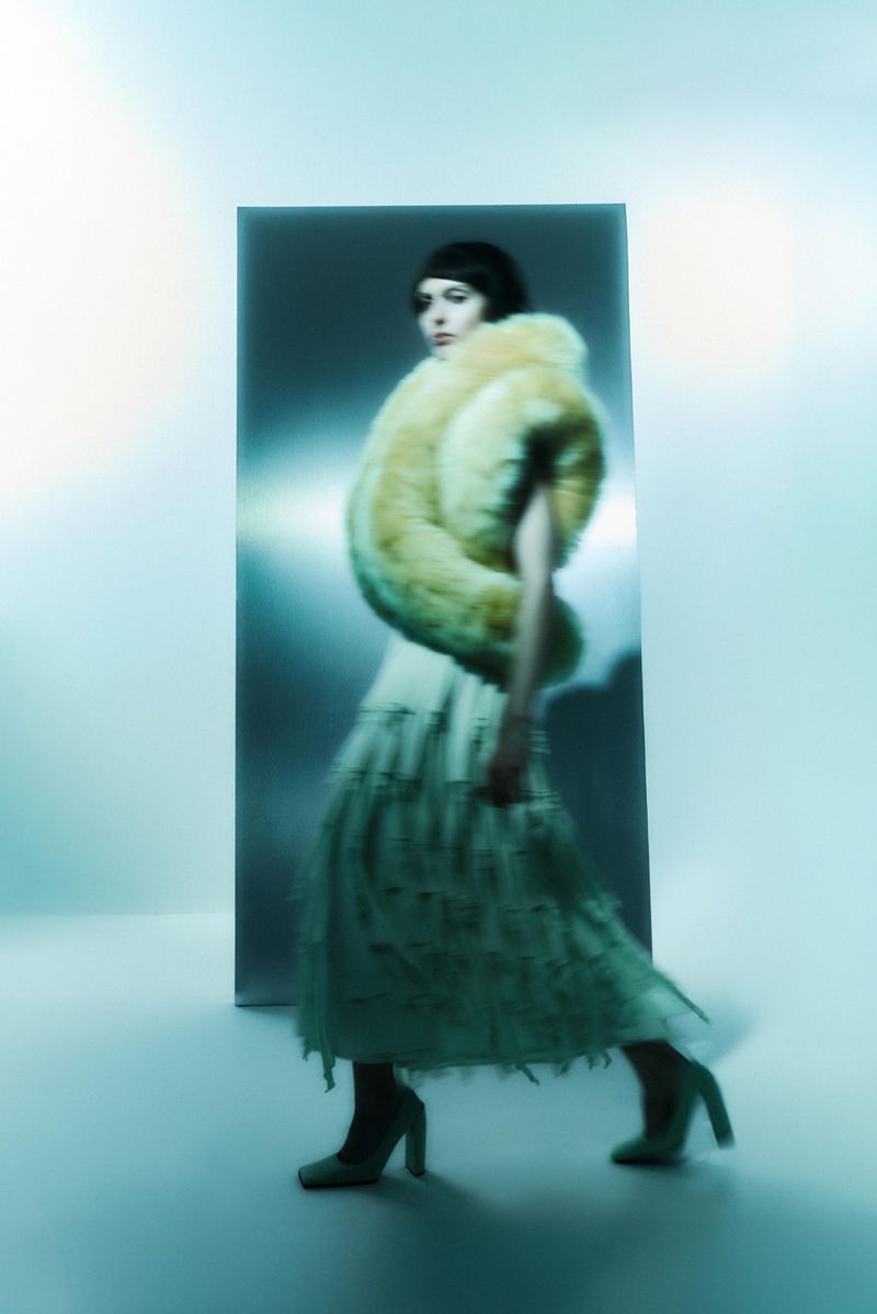 A woman in a cream dress and faux fur stole walks past a rectangular mirror in a brightly lit room with an aqua and cream color palette.