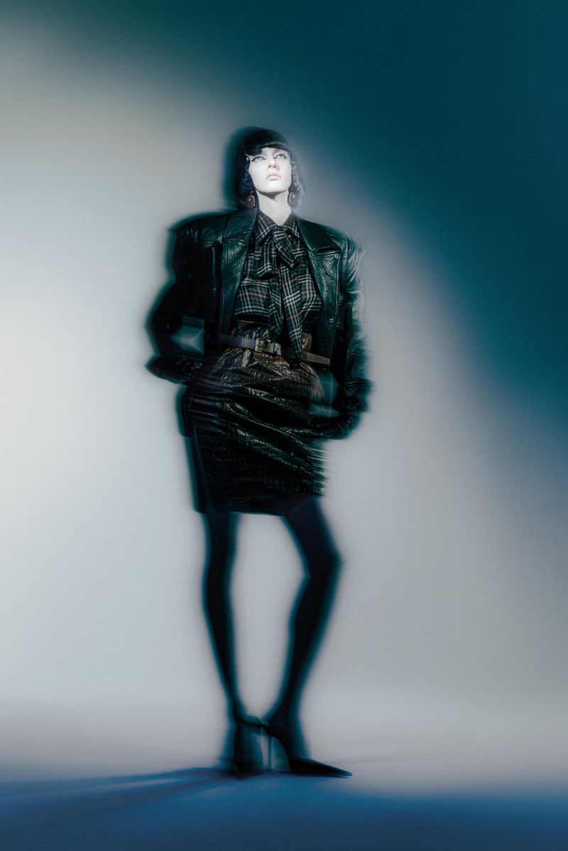 A woman poses in a black leather jacket, skirt, and tights against a gray gradient background in a fashion photography studio shot.