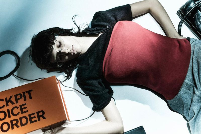 Fashion shot of a woman lying down with an orange Cockpit Voice Recorder box, wearing rimless glasses and a black top in a studio.