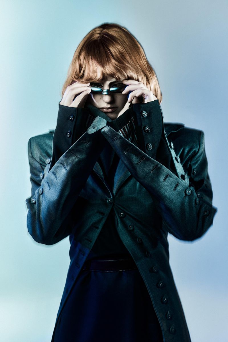 Stylish woman in a futuristic fashion pose, wearing a structured jacket and unique eyewear against a graduated blue backdrop.