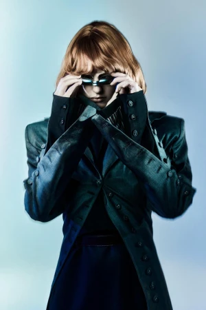 Stylish woman in a futuristic fashion pose, wearing a structured jacket and unique eyewear against a graduated blue backdrop.