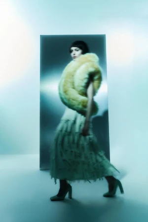 Model walking past a metallic panel wearing a voluminous yellow fur stole and a fringed skirt against a pale blue background.