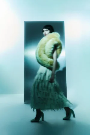 Model walking past a metallic panel wearing a voluminous yellow fur stole and a fringed skirt against a pale blue background.