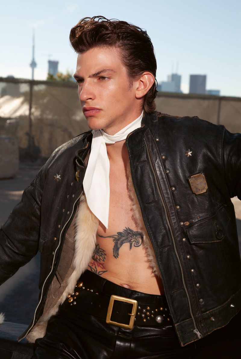 Portrait of adult male in black leather jacket, white neck scarf and tattoos against a urban background with city skyscape.