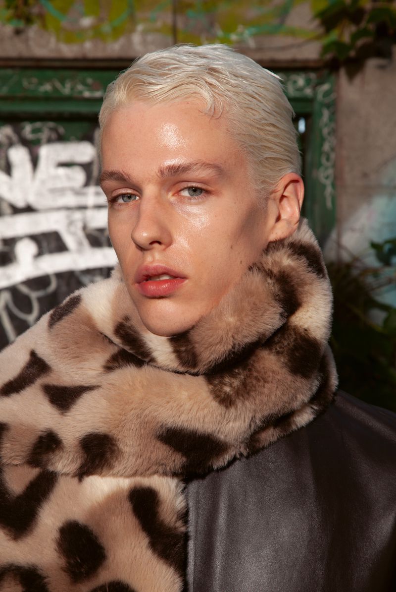 Close-up of a young adult man with blond hair and a fur scarf, posing outdoors against a graffiti-covered background.