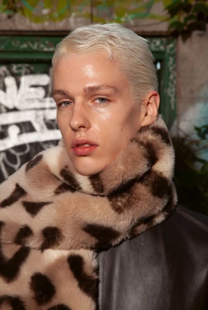 Close-up of a young adult man with blond hair and a fur scarf, posing outdoors against a graffiti-covered background.