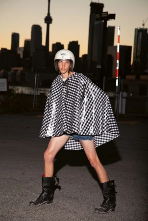 Man posing in black and white checkered poncho and helmet in urban setting with city skyline, and black buckle boots.